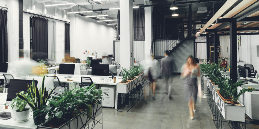Open-office workspace with rows of desks and computers, lush potted plants, and blurred employees walking.