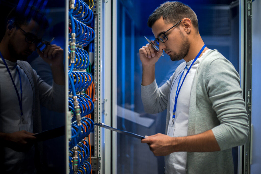 Tech worker in a data center inspecting blue network cables inside a server rack, holding a tablet and wearing a blue lanyard.