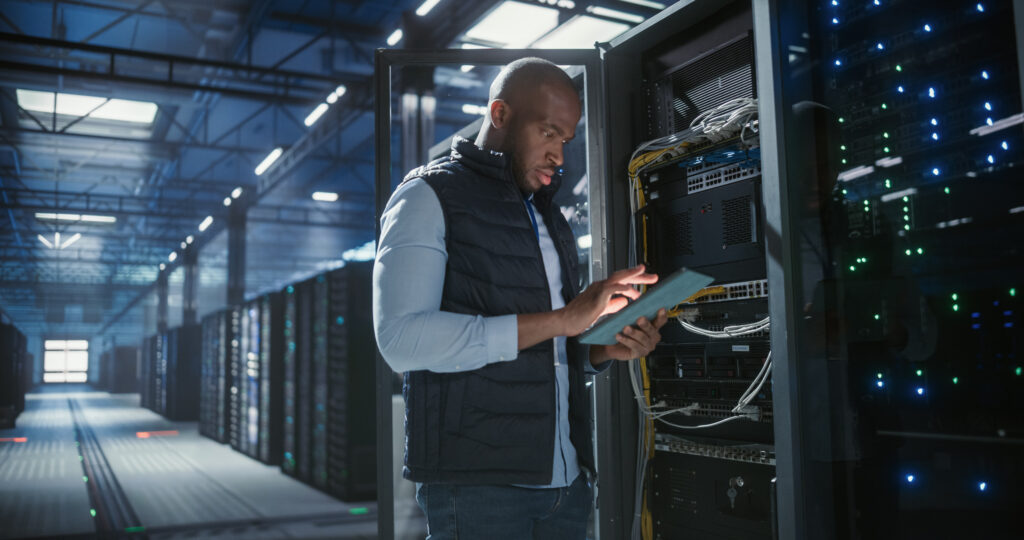 IT technician inspects a server rack in a data center while using a tablet to check connections and status.