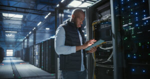 IT technician inspects a server rack in a data center while using a tablet to check connections and status.