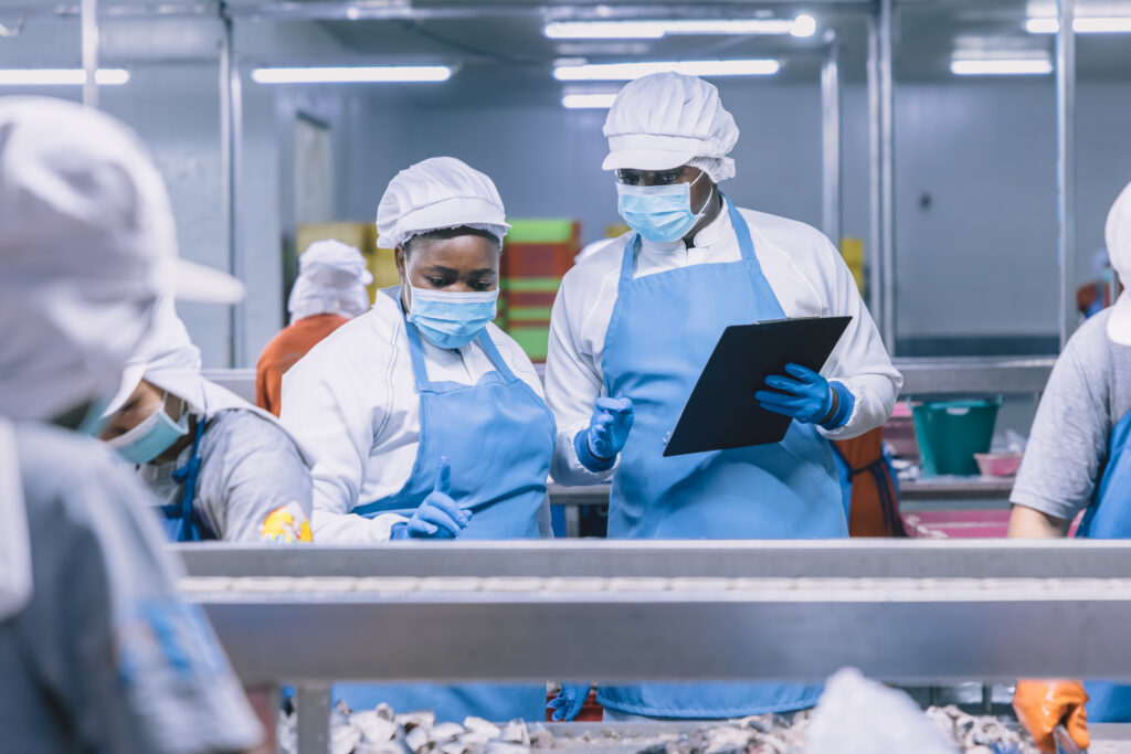 Workers in blue protective aprons and masks inspect materials on a production line in a clean industrial setting.