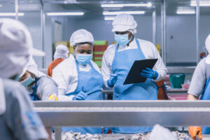 Workers in blue protective aprons and masks inspect materials on a production line in a clean industrial setting.